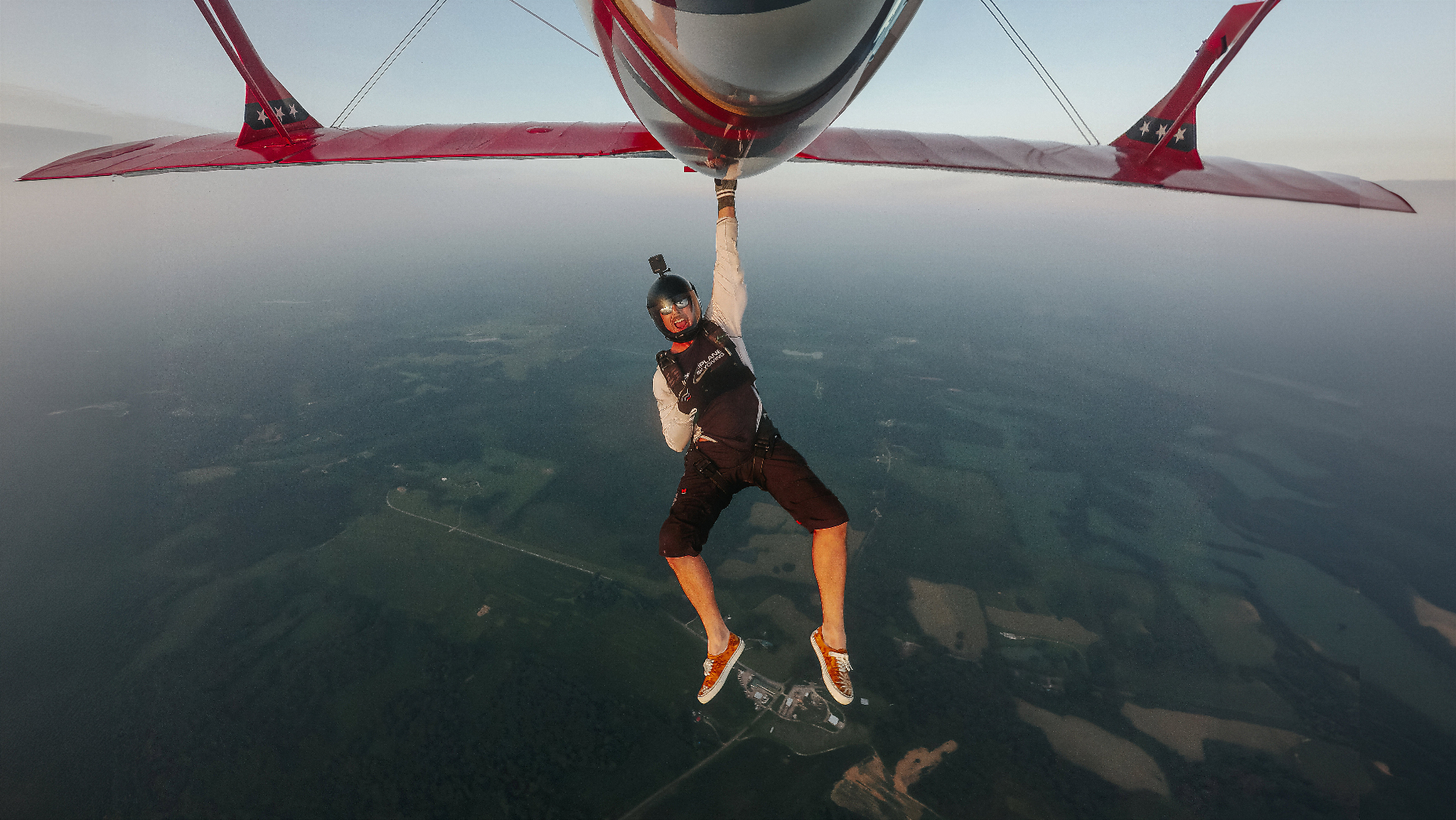 Skydiver hanging from the wing of a red Pitts S2B biplane high above the countryside